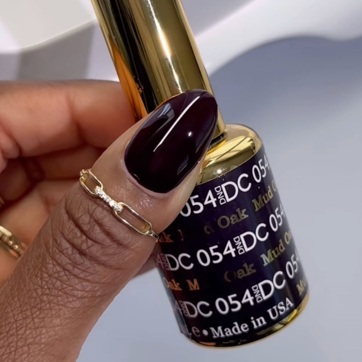 Hand with dark maroon-burgundy nail holding DC #054 gel polish bottle