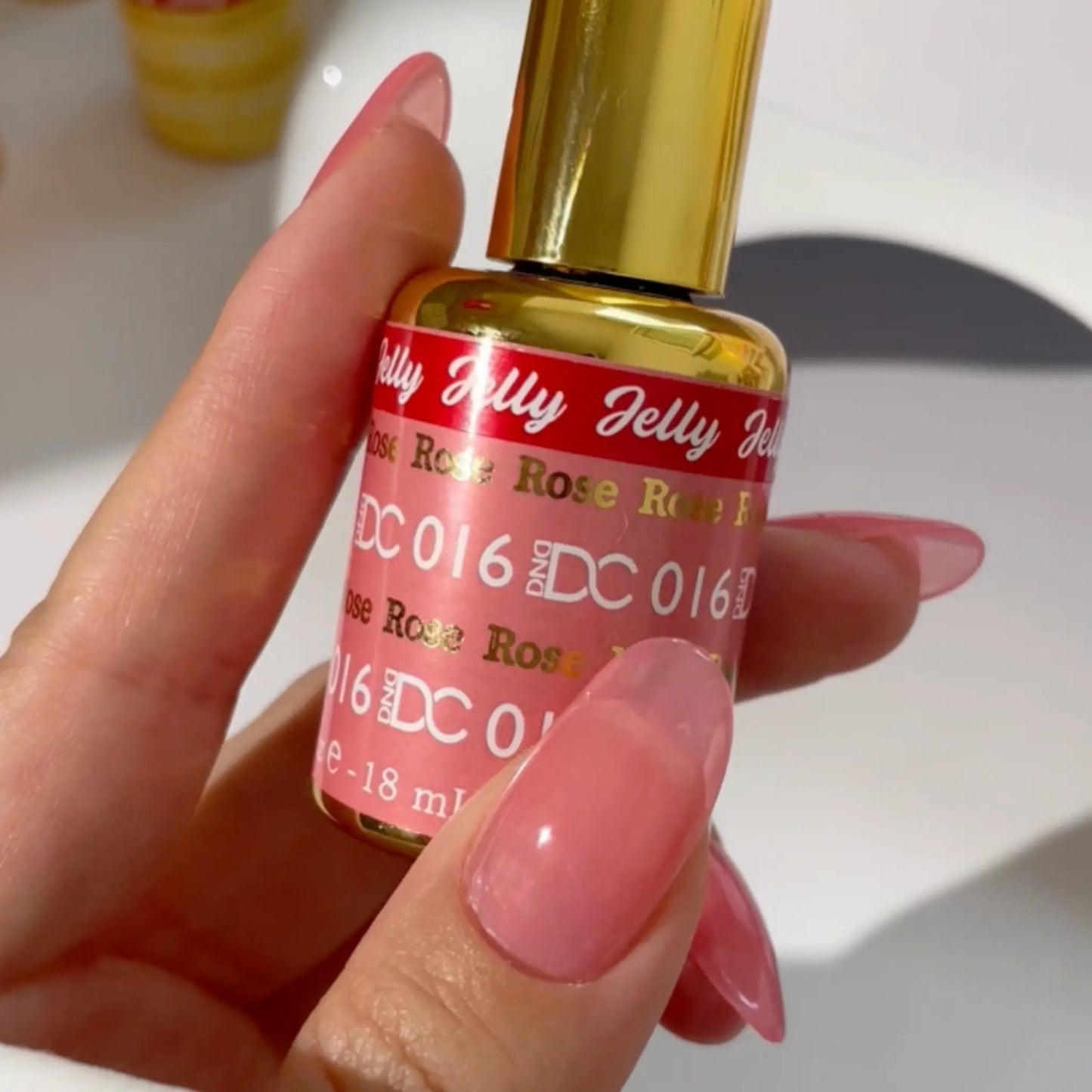 Hand holding a gold-capped DC Jelly #016 Rose gel polish bottle, nails in soft pink