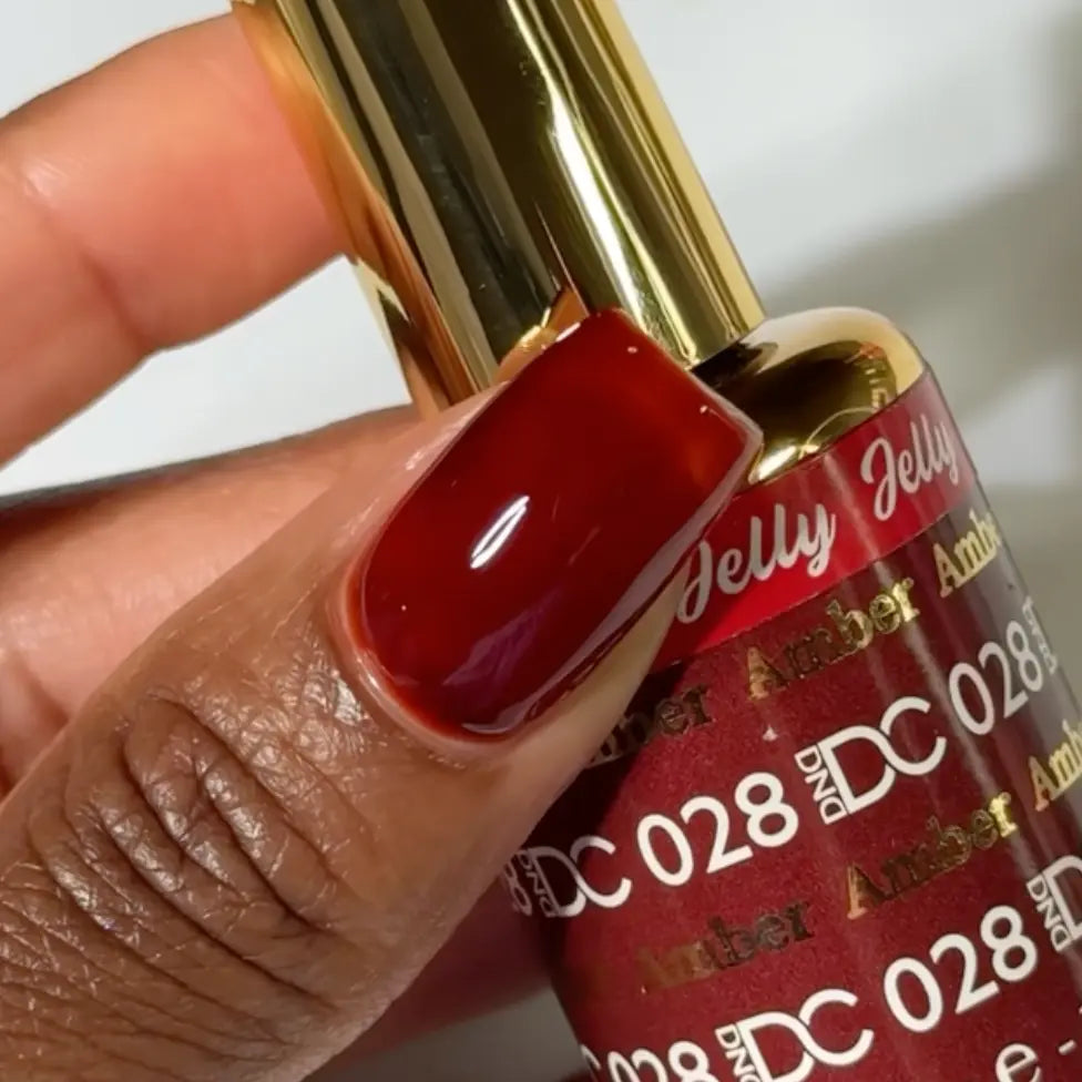Finger with a medium square nail wearing DC Jelly Amber golden brown translucent jelly nail polish, holding a bottle of DND nail polish