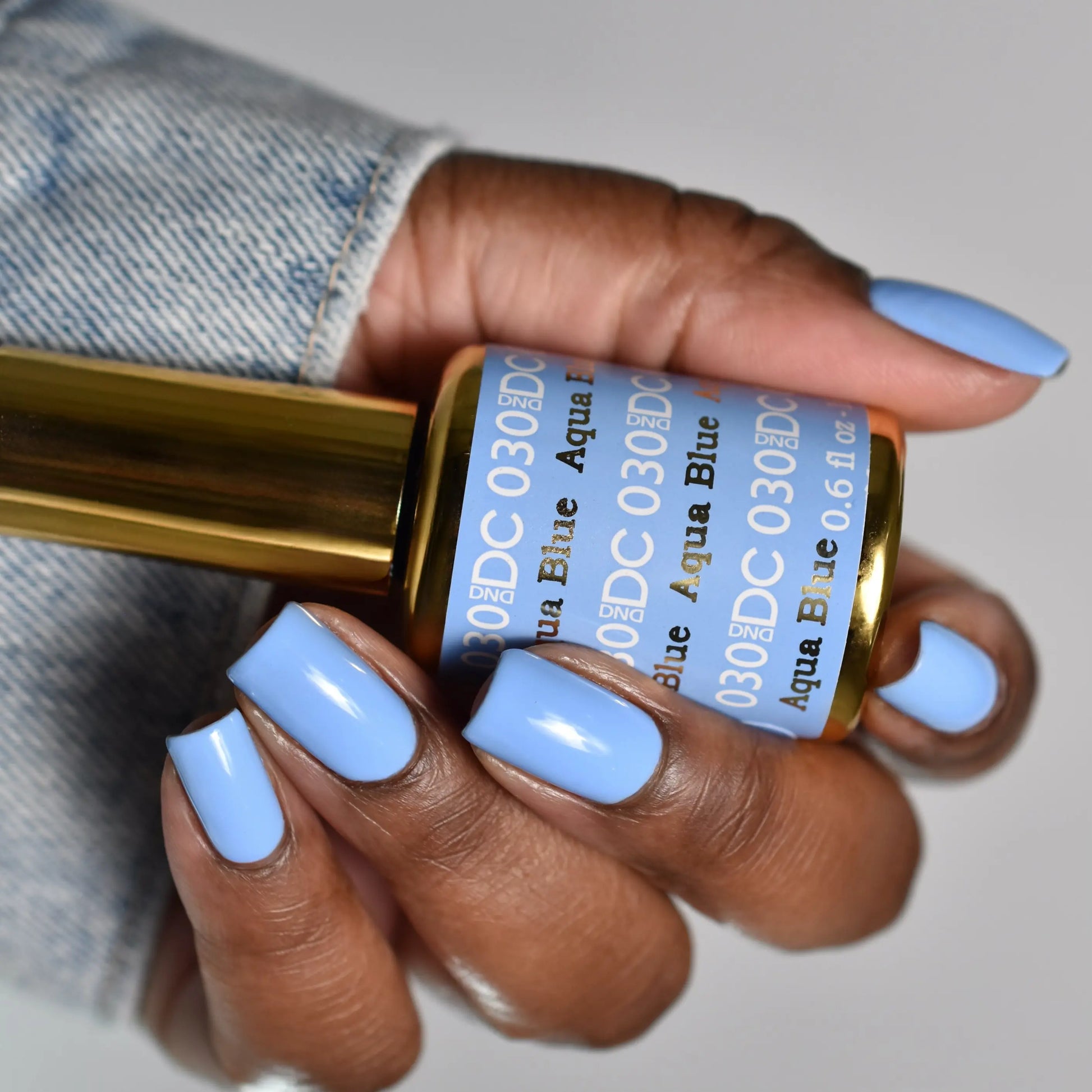 Hand with light blue manicured nails holding a DC Aqua Blue #030 gel nail polish bottle with gold cap