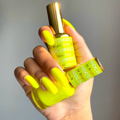 Hand with bright neon yellow manicured nails holding a bottle of DC Shine Bright #258 gel nail polish, fluorescent sunny neon yellow