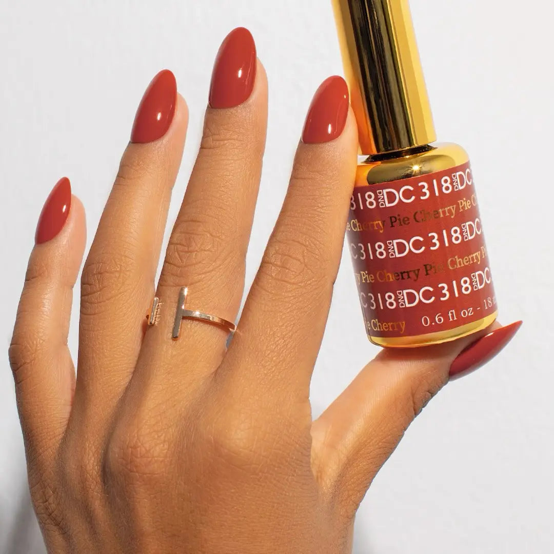 Hand with long glossy nails in DC Cherry Pie #318 gel nail polish, burnt warm autumn red, holding matching bottle