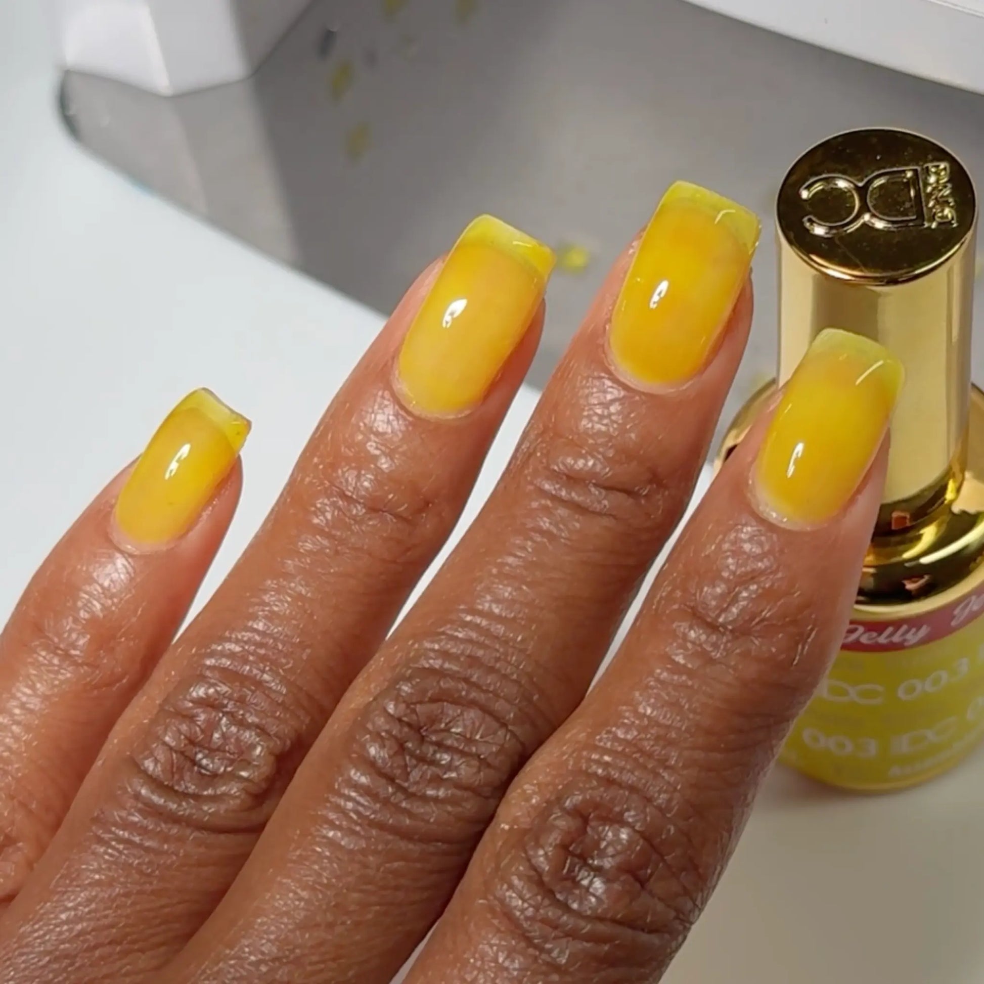 Hand with glossy DC Jelly Yuzu yellow jelly manicure next to the bottle
