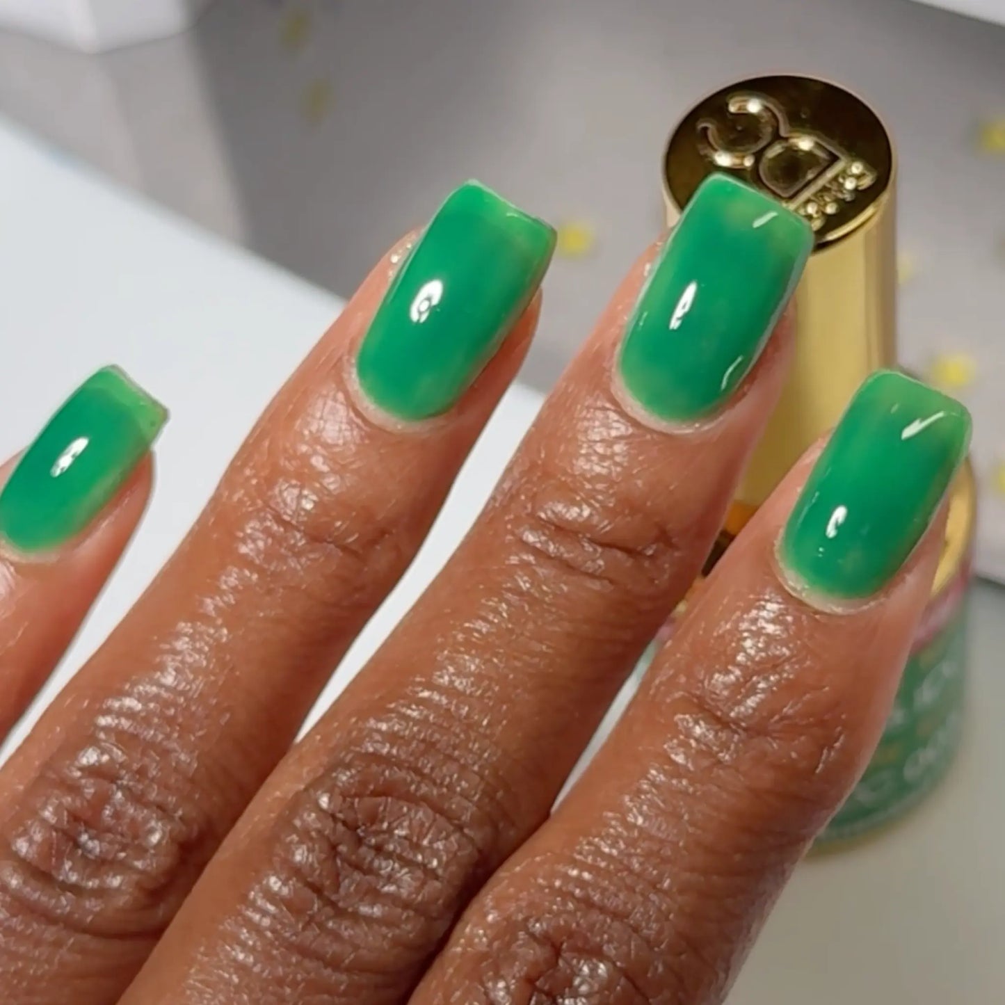 Close-up of a hand with square-shaped nails wearing DC Jelly Kelly emerald green jelly nail polish, with a bottle of DND nail polish nearby