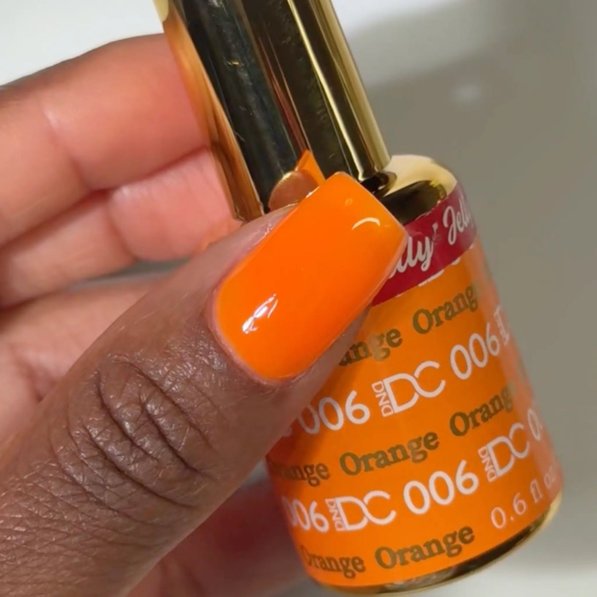 Close-up of a fingernail painted with DC Jelly Orange true orange jelly nail polish, holding a bottle of DND nail polish