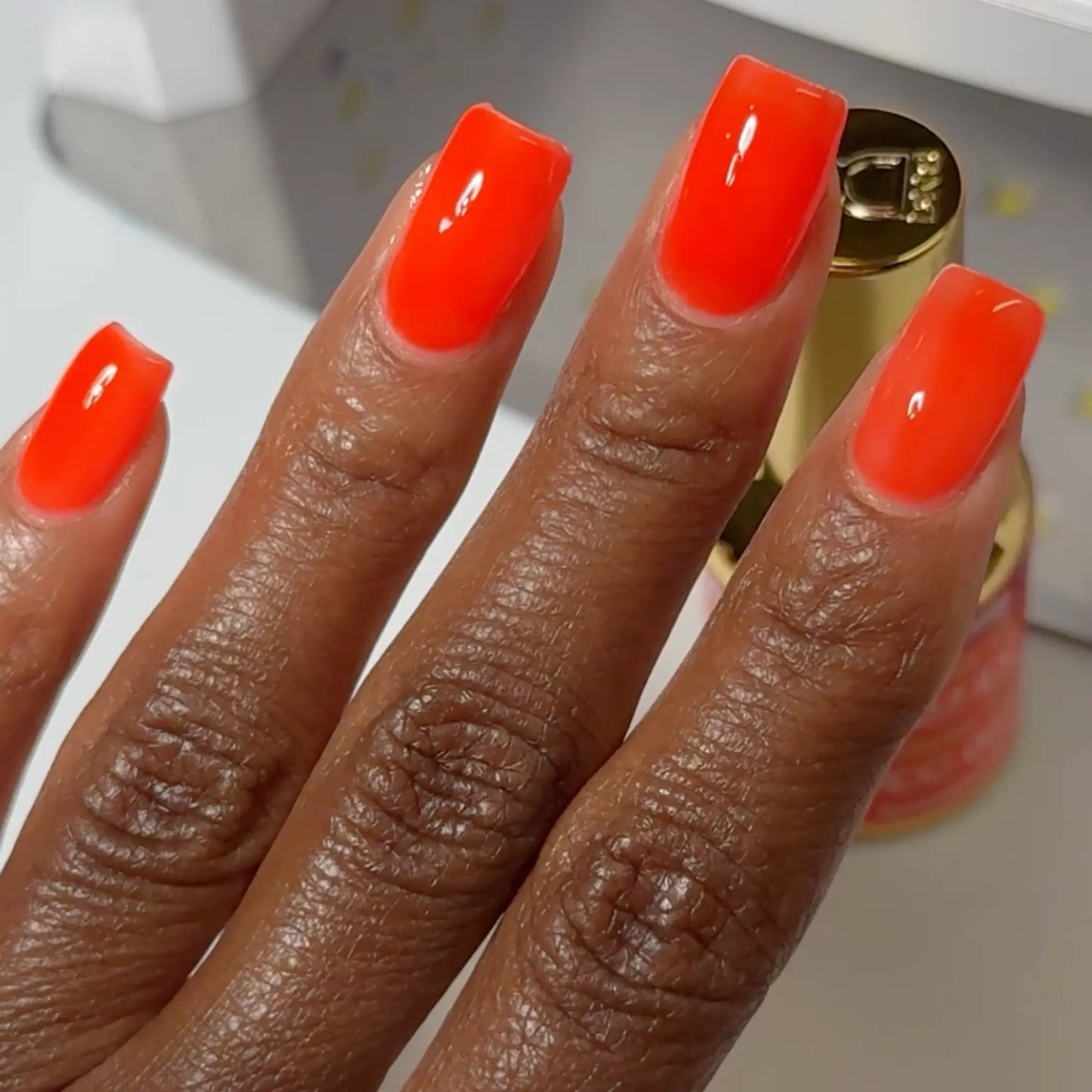 Close-up of a hand with long square-shaped nails wearing DC Jelly Citrus fiery orange jelly nail polish, with a bottle of DND nail polish in the background