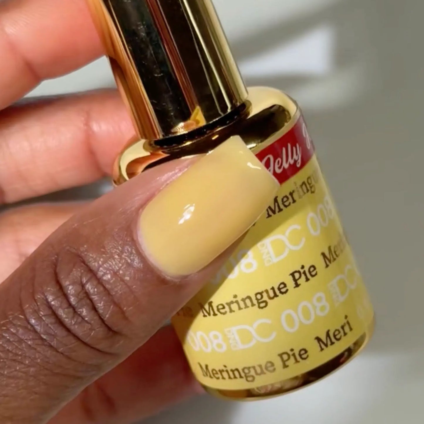 Close-up of a fingernail painted with DC Jelly Merengue Pie pale yellow jelly nail polish, holding a bottle of DND nail polish