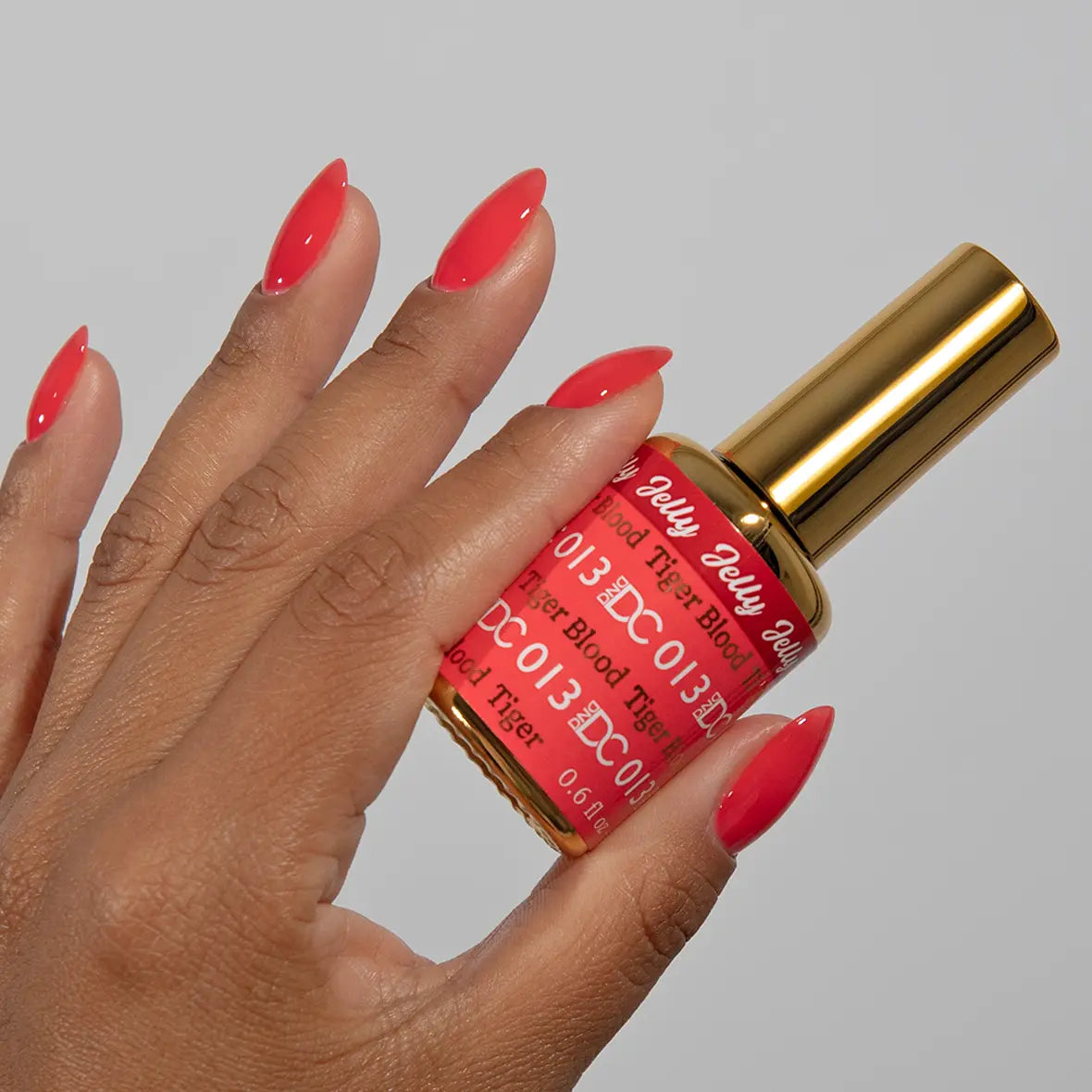 Hand with long almond nails wearing DC Jelly Tiger Blood bright coral-red jelly nail polish, holding a bottle of DND nail polish