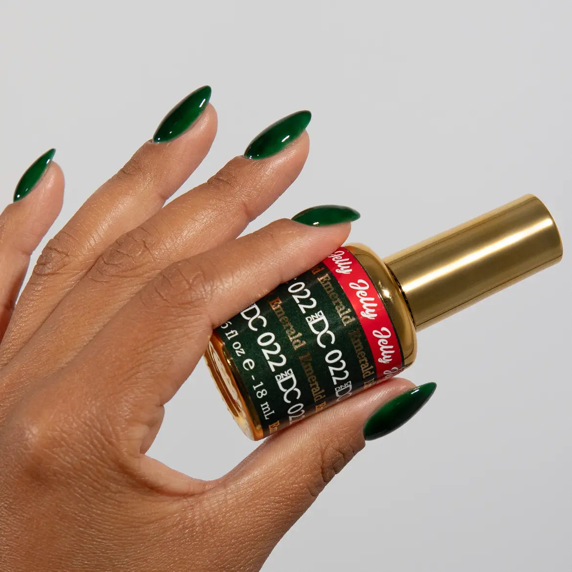 Hand with long almond nails wearing DC Jelly Emerald deep green jelly nail polish, holding a bottle of DND nail polish