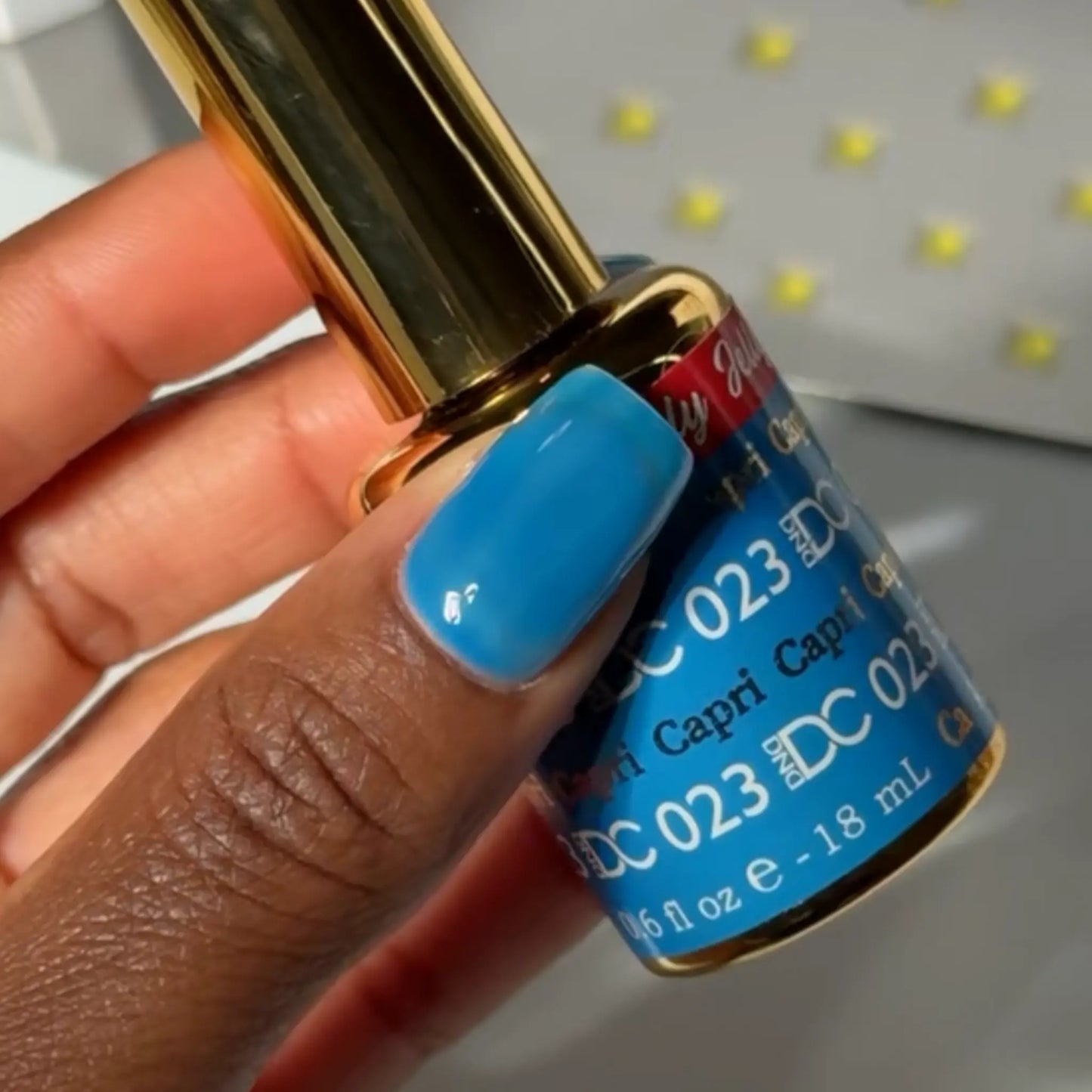 Finger with a square nail wearing DC Jelly Capri bright azure blue jelly nail polish, holding a bottle of DND nail polish