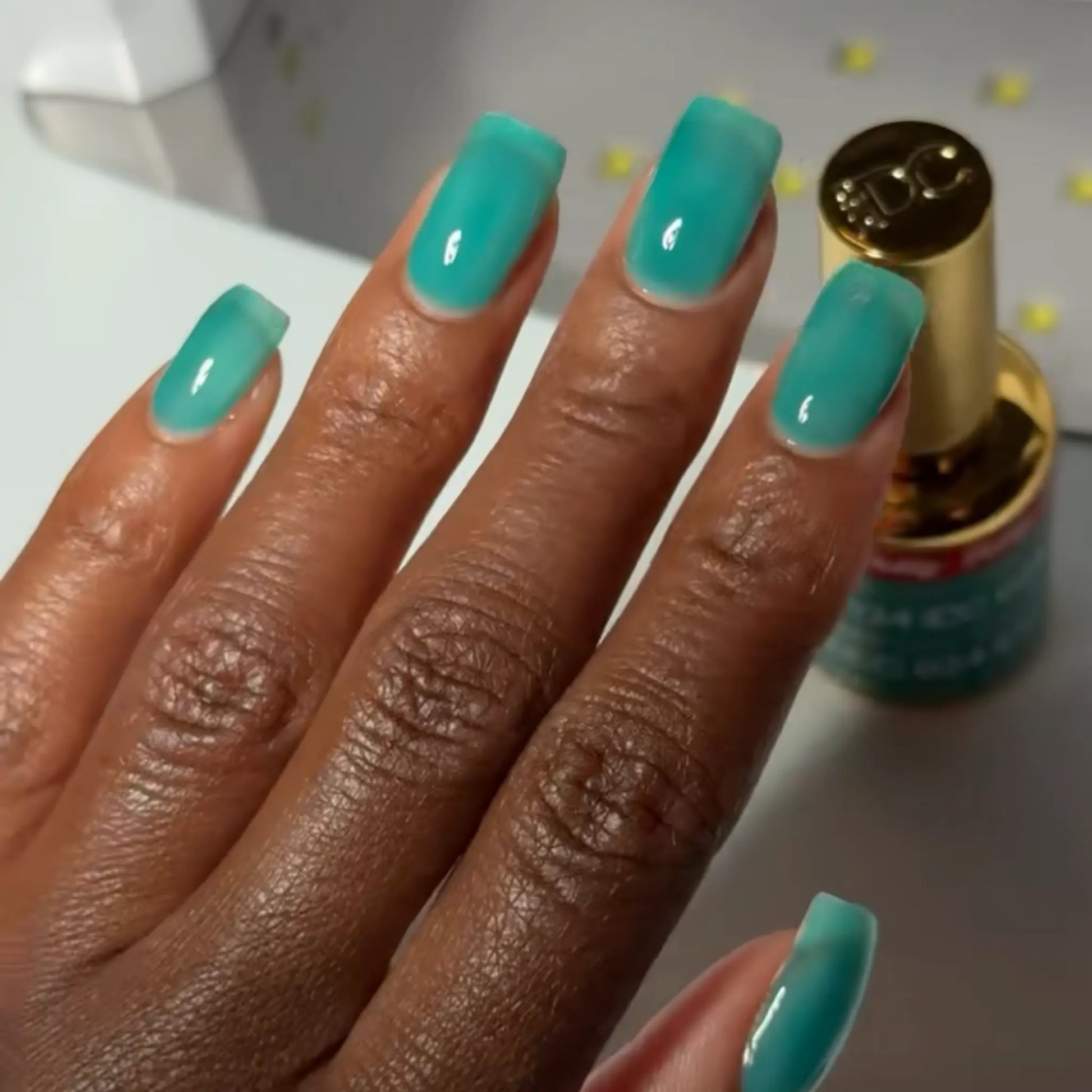 Hand with square nails wearing DC Jelly Jade turquoise green-blue jelly nail polish, next to a bottle of DND nail polish