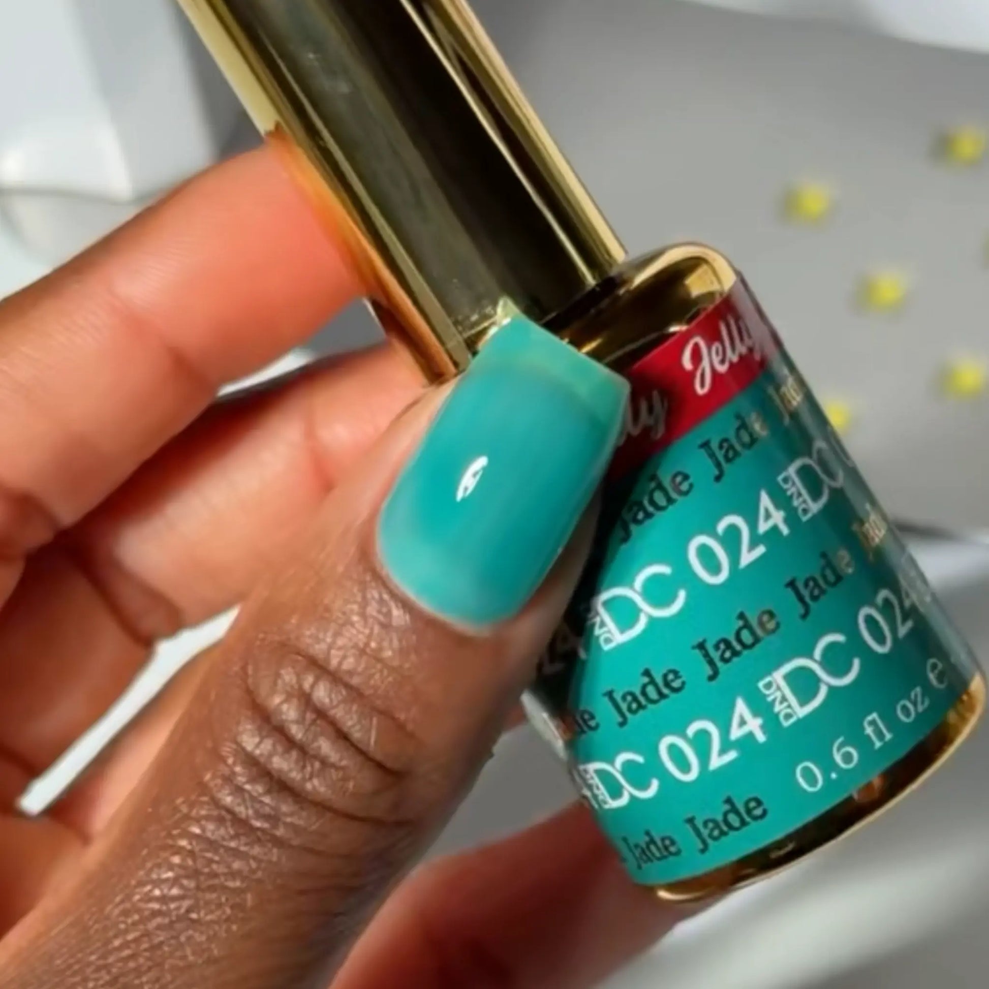 Finger with a square nail wearing DC Jelly Jade turquoise green-blue jelly nail polish, holding a bottle of DND nail polish