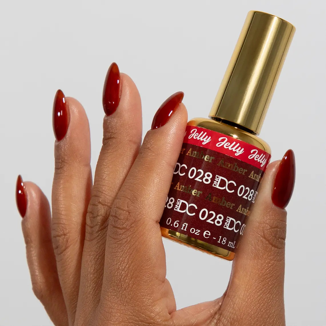 Hand with long almond nails wearing DC Jelly Amber deep amber brown-red jelly nail polish, holding a bottle of DND nail polish