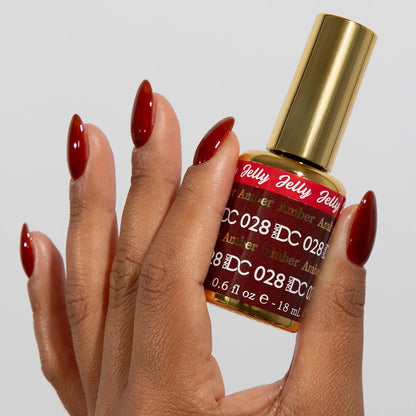 Hand with long almond nails wearing DC Jelly Amber deep amber brown-red jelly nail polish, holding a bottle of DND nail polish