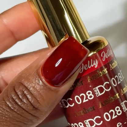 Finger with a medium square nail wearing DC Jelly Amber golden brown translucent jelly nail polish, holding a bottle of DND nail polish