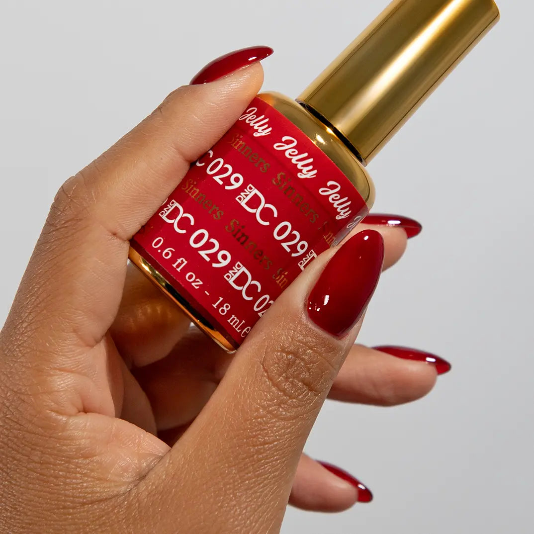 Hand with long stiletto nails wearing DC Jelly Sinners deep ruby red jelly nail polish with orange undertones, holding a bottle of DND nail polish