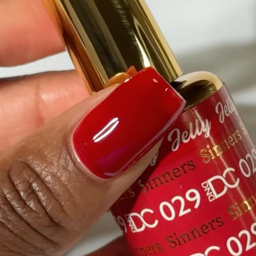 Finger with a square nail wearing DC Jelly Sinners deep ruby red jelly nail polish with orange undertones, holding a bottle of DND nail polish