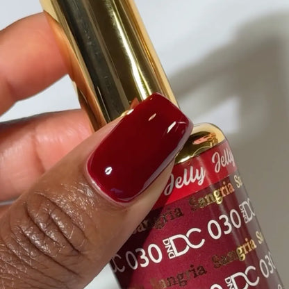 Finger with a square nail wearing DC Jelly Sangria deep red jelly nail polish, holding a bottle of DND nail polish