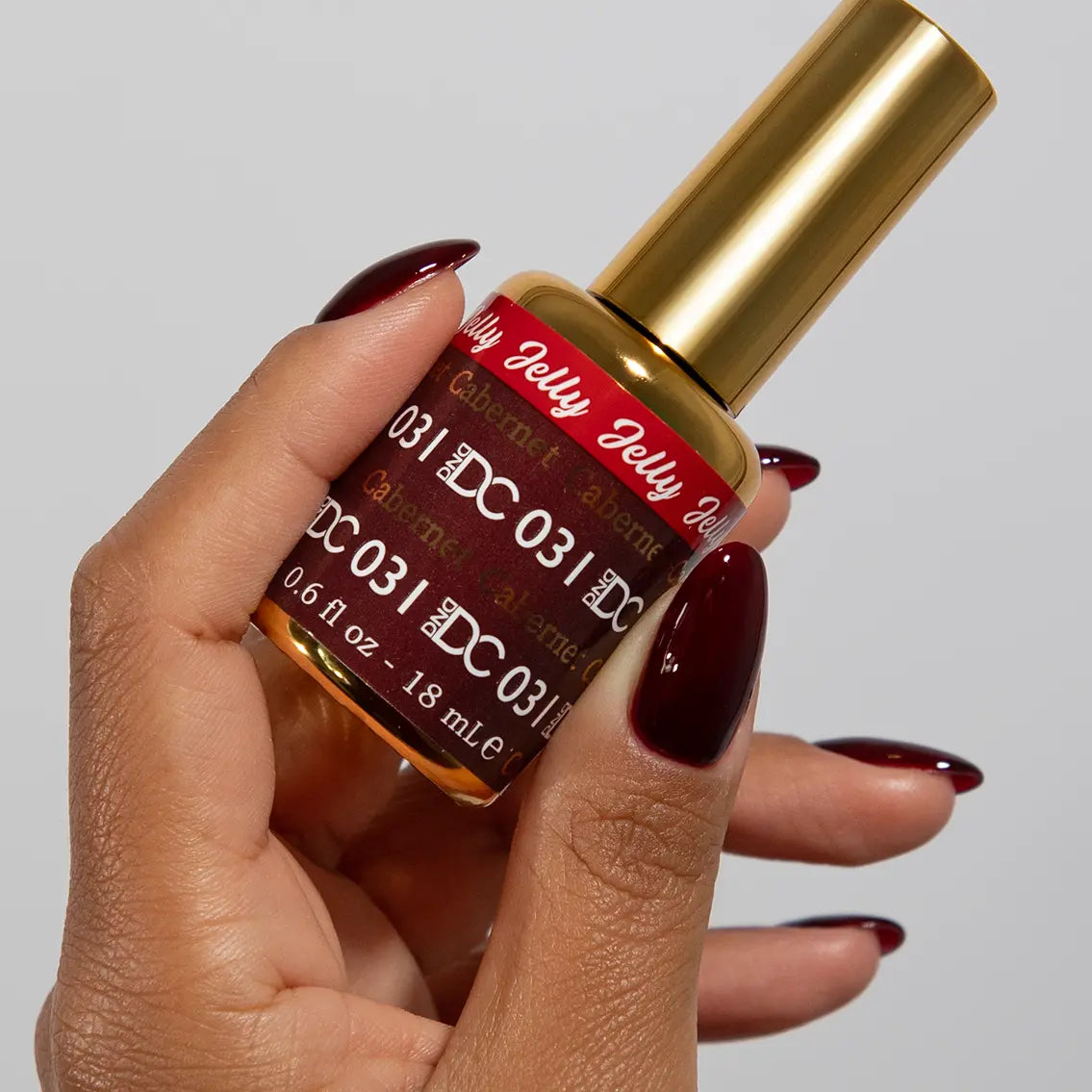 Hand with long almond nails wearing DC Jelly Cabernet scarlet red jelly nail polish, holding a bottle of DND nail polish