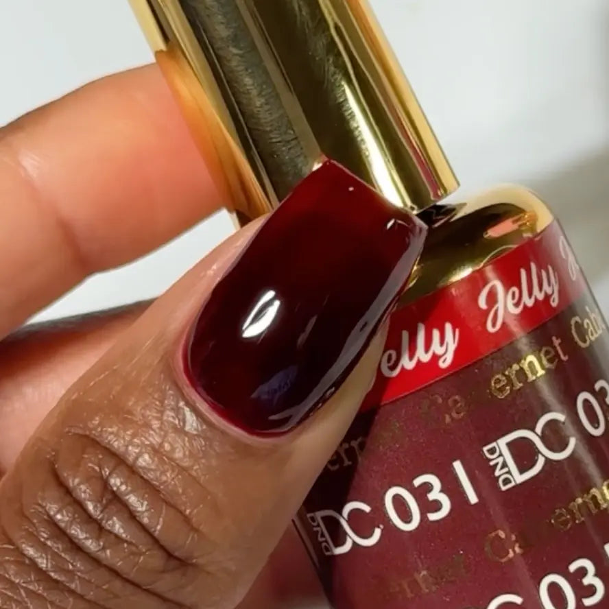 Finger with a square nail wearing DC Jelly Cabernet deep burgundy wine jelly nail polish, holding a bottle of DND nail polish