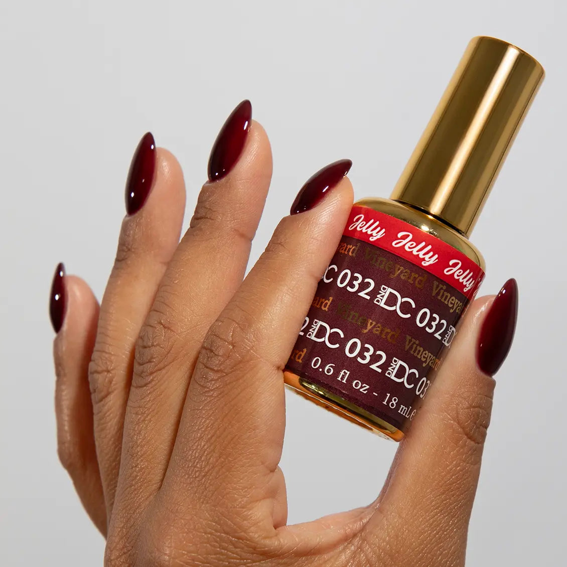 Hand with long almond nails wearing DC Jelly Vineyard deep cinnamon red jelly nail polish with a purple flush, holding a bottle of DND nail polish