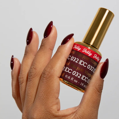 Hand with long almond nails wearing DC Jelly Vineyard deep cinnamon red jelly nail polish with a purple flush, holding a bottle of DND nail polish