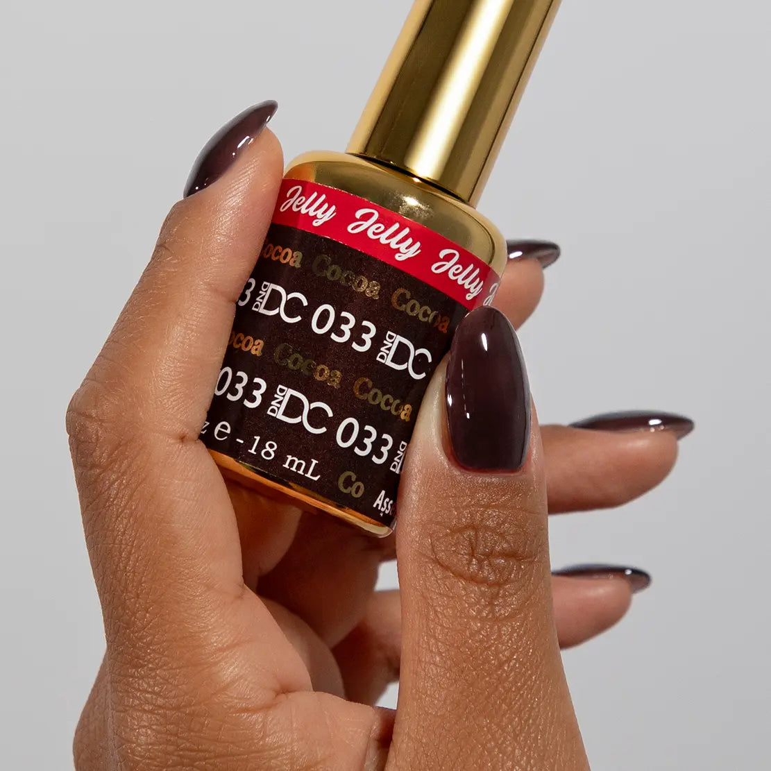 Hand with long almond nails wearing DC Jelly Cocoa muted soft brown jelly nail polish, holding a bottle of DND nail polish