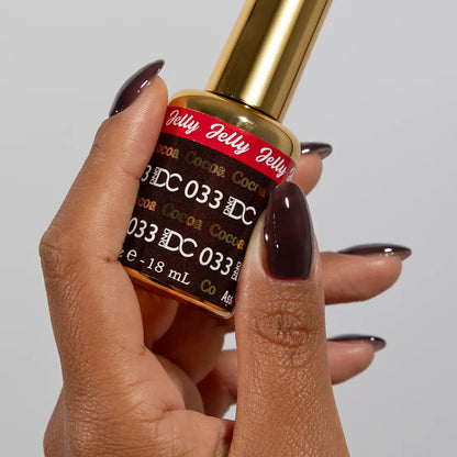 Hand with long almond nails wearing DC Jelly Cocoa muted soft brown jelly nail polish, holding a bottle of DND nail polish
