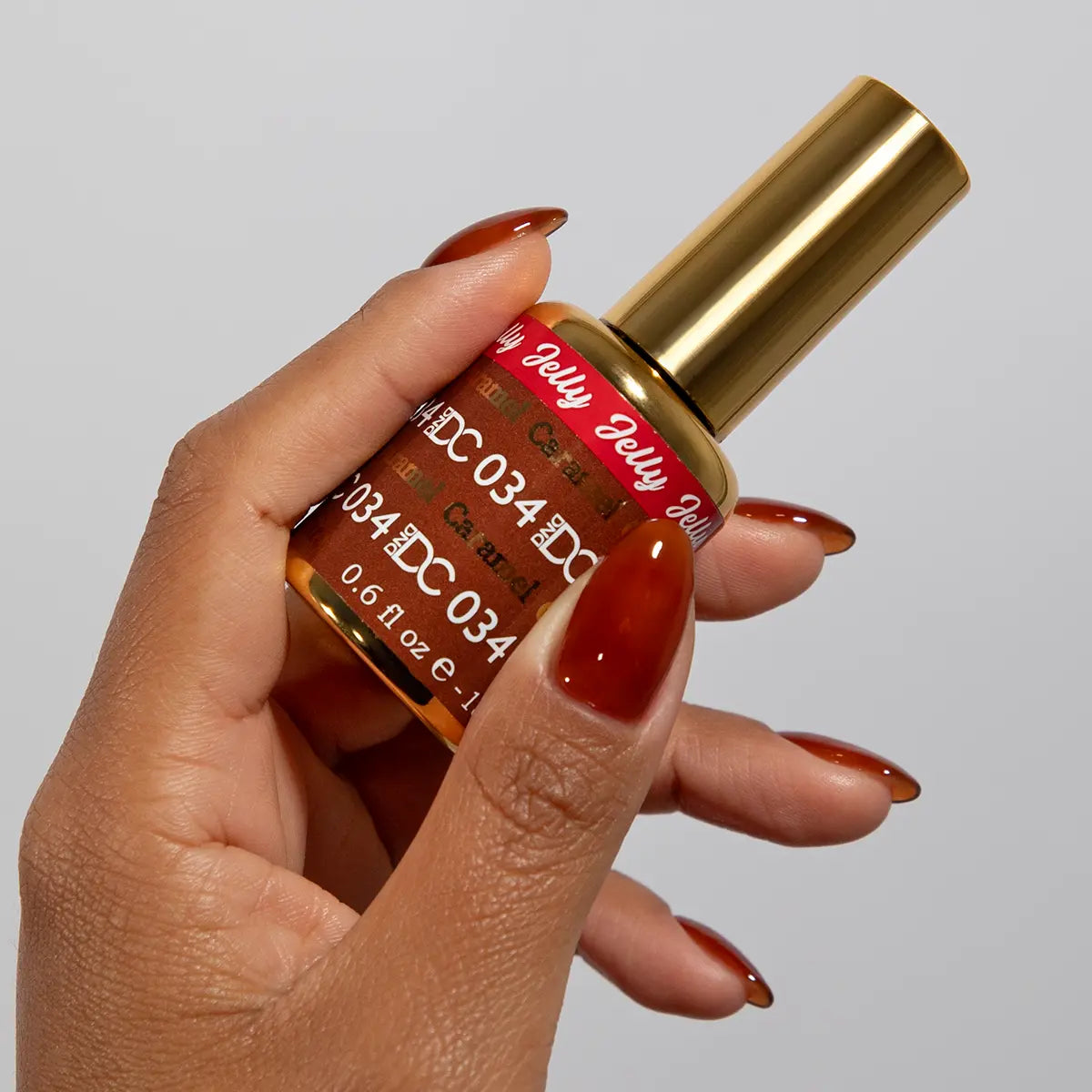 Hand with long stiletto nails wearing DC Jelly Caramel burnt-honey orange-brown jelly nail polish, holding a bottle of DND nail polish