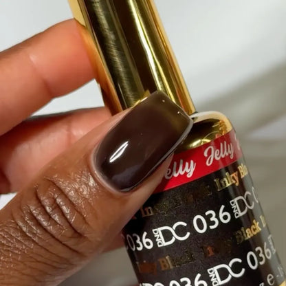 Finger with a square nail wearing DC Jelly Inky Black translucent black-brown jelly nail polish, holding a bottle of DND nail polish