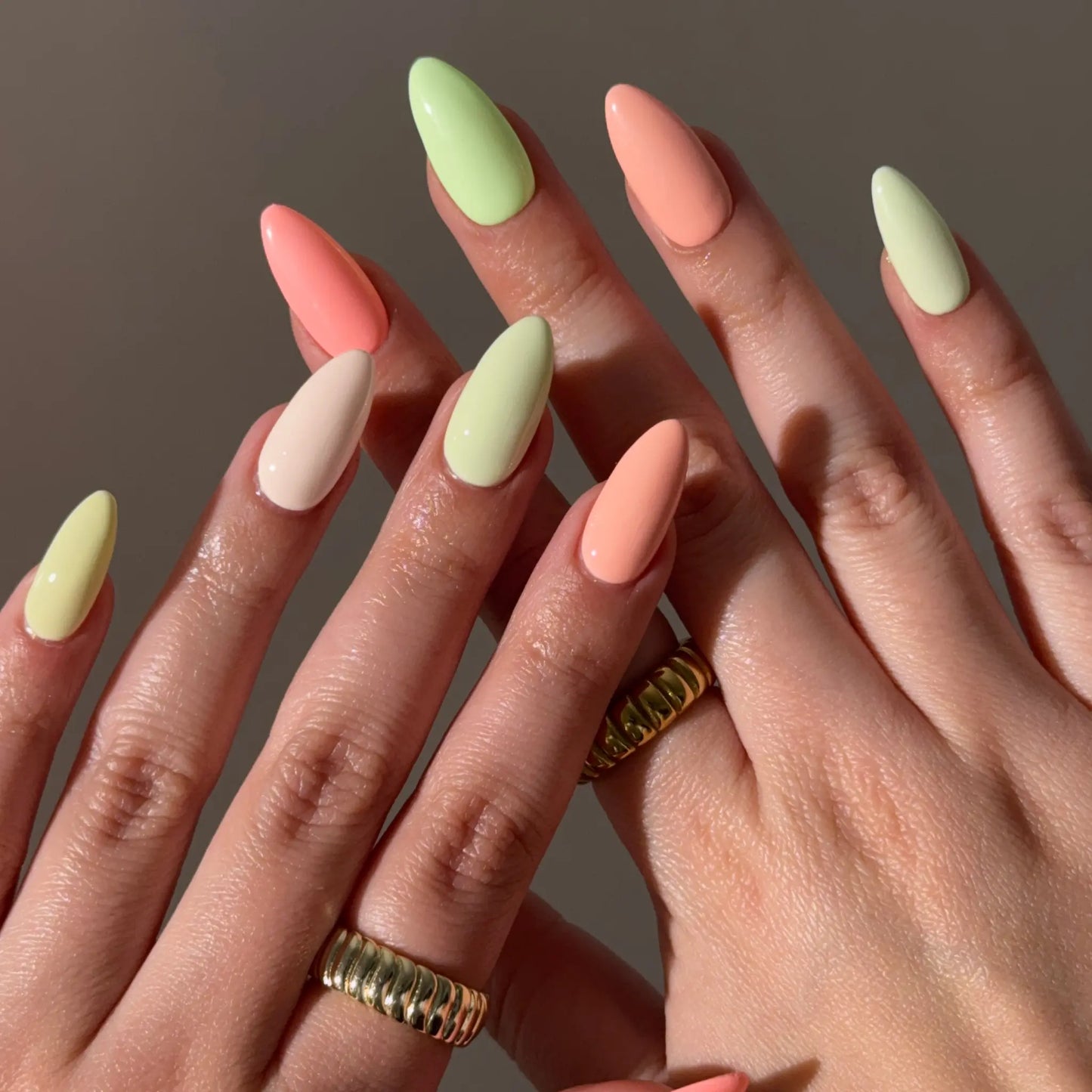 Close-up of hands with pastel citrus nail colors and gold rings on a neutral background