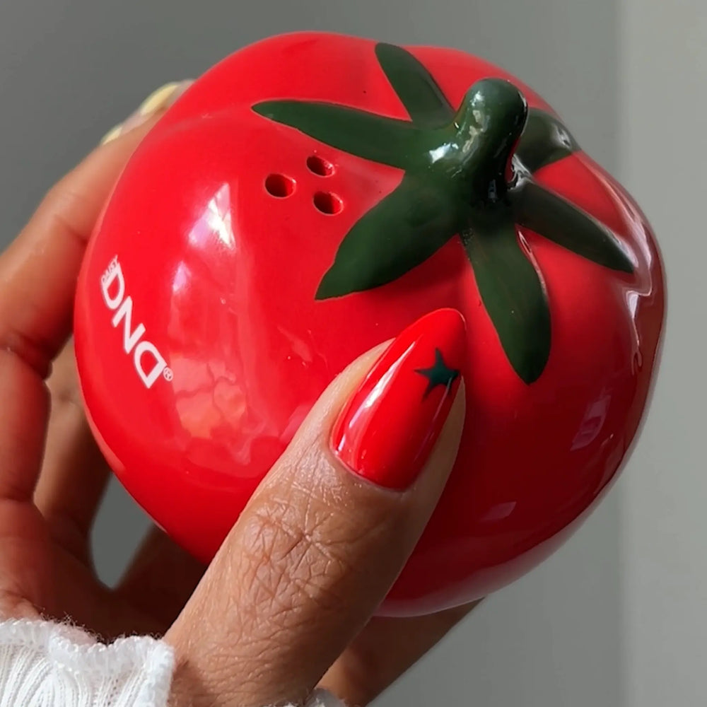 Hand with a glossy red nail and dark green star nail art resting on a red tomato-shaped DND accessory, DND Yes Chef #006 Scorching