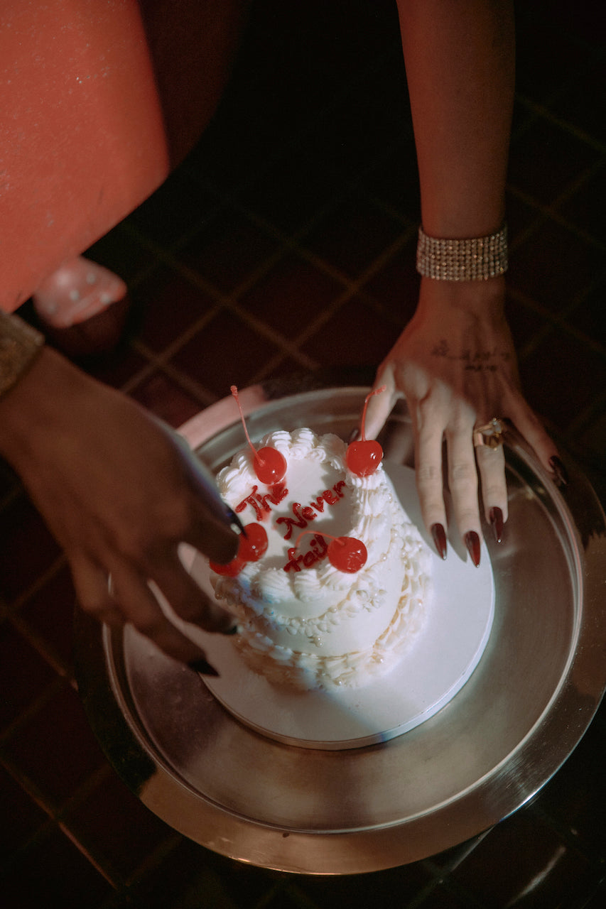 Hands with dark red nails decorating a cake with cherries and "The Never Fail" piped icing text, for the DND DEL gel polish collection