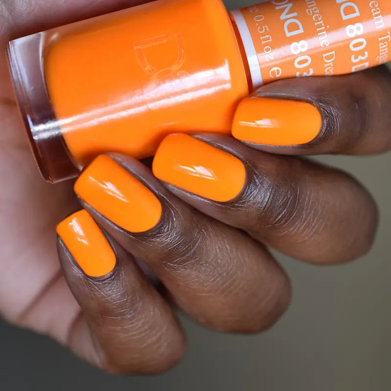 Hand with medium square/oval nails wearing tangerine dreams vibrant orange nail polish, holding a matching bottle of DND nail polish